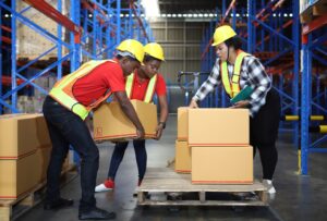 A group of 3 warehouse workers wearing yellow protective helmets handle pallets.