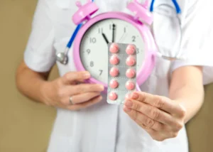 A healthcare professional holds up a blister pack of round pink pills in front of a clock. 
